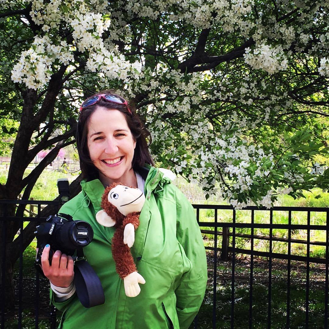 Smiling woman wearing green jacket, holding camera with a stuffed monkey (child's toy) peeking out from inside the jacket.