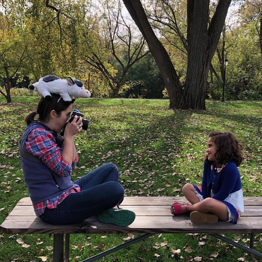 Woman sitting on picnic table, holding a camera and balancing a stuffed cow on her head, while photographing a young girl who is also sitting on the picnic table across from her.